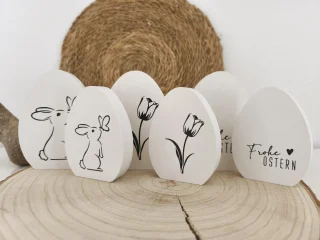 Weiße Osterei-Dekoration aus Holz mit Hasen-, Tulpen- und Oster-Schriftmotiven auf einer natürlichen Holzscheibe.